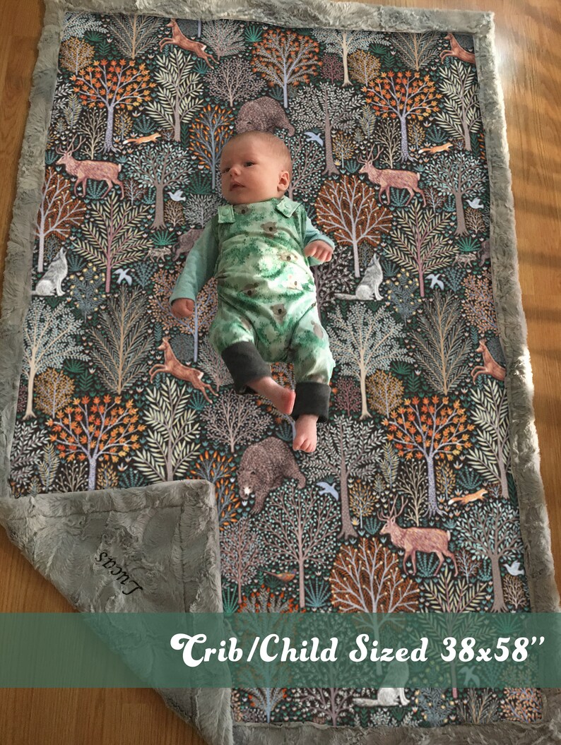 May include: A crib-sized baby blanket with a woodland print featuring trees, animals, and birds. The blanket is gray with a soft, fuzzy texture and has a white border. The blanket is 38 inches by 58 inches.