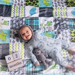 以下が含まれることがあります： 青いクマ柄のロンパースと灰色のニット帽をかぶった赤ちゃんが、グレーとグリーンのヘリンボーン柄のマルチカラーの毛布の上に寝ています。赤ちゃんは灰色の編みぐるみテディベアを持っています。毛布の上に「02ヶ月」と書かれた木製のブロックがあります。