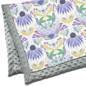 May include: A soft, grey baby blanket with a white background and a colourful floral pattern featuring hummingbirds, butterflies, and bees. The blanket has a textured, grey border.