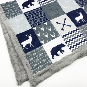 Puede incluir: Una manta para bebé azul marino y gris con un diseño de patchwork que presenta ciervos, osos y flechas blancas. La manta tiene un borde gris suave y mullido.