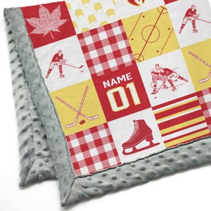 May include: A red, white and yellow patchwork baby blanket with a hockey theme. The blanket features a hockey player, a hockey stick, a hockey skate and a hockey puck. The blanket also has a red and white checked pattern, a yellow and red striped pattern and a maple leaf. The blanket is personalised with the text "NAME 01".