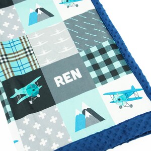以下が含まれることがあります： 青い飛行機、山、白い文字で「REN」と書かれた、青とグレーのパッチワークのベビーブランケット。ブランケットは青いミンクのトリムが付いています。