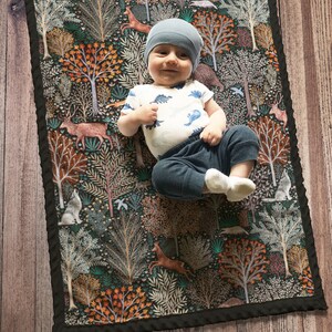 May include: A baby blanket with a woodland theme. The blanket features a dark background with colorful trees, animals, and flowers. The blanket is 28 inches by 38 inches.