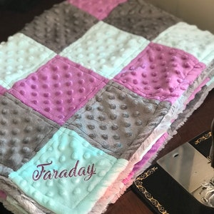 Peut inclure: Une couverture de bébé douce et moelleuse avec un motif patchwork dans des tons de gris, blanc et violet. La couverture est personnalisée avec le nom "Faraday" brodé en cursive sur un carré bleu clair.