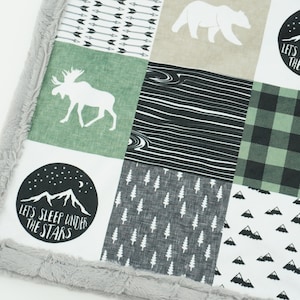 Peut inclure: Une couverture bébé en patchwork vert et noir avec un élan, un ours et des montagnes. La couverture a une bordure douce et duveteuse et présente le texte "Let's Sleep Under The Stars".