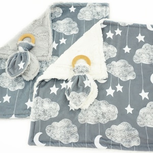 Peut inclure: Une couverture bébé grise avec un motif de nuages et d'étoiles blancs. La couverture est faite d'un tissu doux et pelucheux et comprend un hochet assorti avec un anneau en bois.