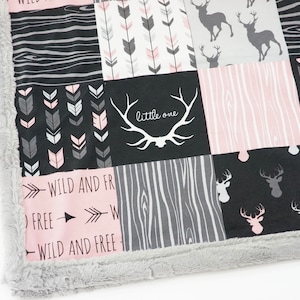 May include: A pink and black baby blanket with a patchwork design featuring deer, arrows, and wood grain patterns. The blanket has a grey faux fur border and the text "wild and free" and "little one" is printed on the blanket.