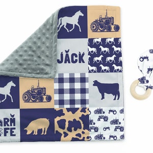 Op de afbeelding: Een blauwe en bruine babydeken met een patchwork-ontwerp met boerderijdieren, tractoren en de naam "JACK". Een houten bijtring met stoffen tractoraccenten is inbegrepen.