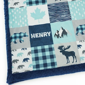 May include: A blue and green patchwork baby blanket featuring a moose, bears, mountains, and a maple leaf design. The blanket has a navy blue border and the name "HENRY" is embroidered in the centre.