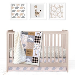 May include: A baby crib with a white crib sheet and a patchwork quilt with a giraffe, elephants, and the words "Wild One" and "One". The crib has a pink and gray elephant print skirt.