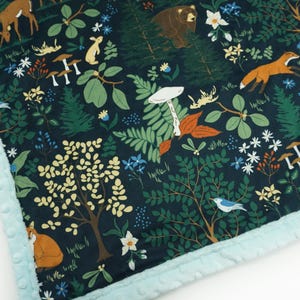 May include: A dark blue fabric with a woodland pattern featuring a variety of animals, plants, and mushrooms. The fabric has a light blue fuzzy trim.