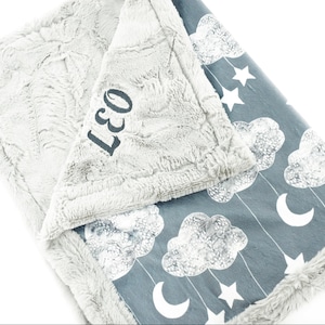Peut inclure: Une couverture pour bébé grise et blanche avec un motif de nuages et d'étoiles. La couverture est personnalisée avec le nom "Leo" en broderie noire.