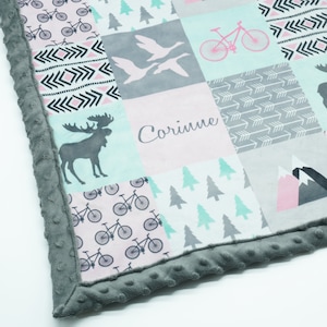 May include: A grey and white baby blanket with a patchwork design. The blanket features a variety of patterns, including bicycles, trees, arrows, and a moose. The blanket is personalised with the name "Corinne".