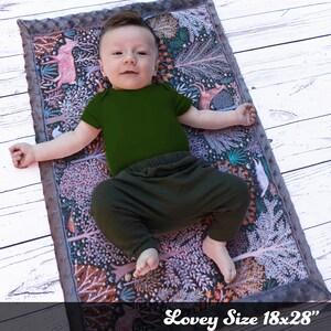 May include: A baby lying on a gray and white patterned baby blanket with a forest scene. The blanket is 18x28 inches and has a soft, plush texture. The baby is wearing a green onesie and black pants.