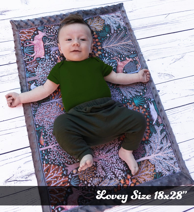 May include: A baby lying on a gray and white patterned baby blanket with a forest scene. The blanket is 18x28 inches and has a soft, plush texture. The baby is wearing a green onesie and black pants.