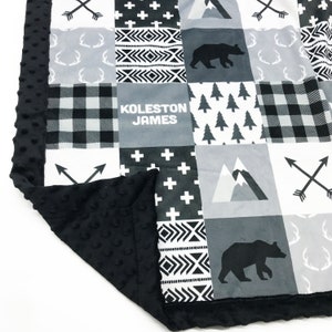 Peut inclure: Une couverture pour bébé noire et blanche avec un motif patchwork. La couverture présente divers motifs, notamment du plaid, des flèches, des montagnes, des arbres et des ours. La couverture est personnalisée avec le nom "Koleston James".