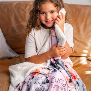 May include: A pink and white floral blanket with a soft, white sherpa lining. The blanket features a personalized name, "Lucy Karmen", in a cursive font. The design includes illustrations of trees and flowers. The blanket is wrapped around a person.