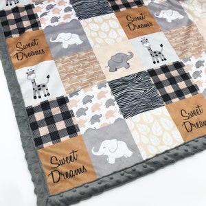 Könnte beinhalten: Eine Patchwork-Decke mit Quadraten in Elefanten-, Giraffen-, Zebramuster-, Pfeil-, Blatt- und Karomustern. Mehrere hellbraune Quadrate zeigen den Text Sweet Dreams in schwarzer Schrift. Die Decke hat einen strukturierten grauen Noppenrand.