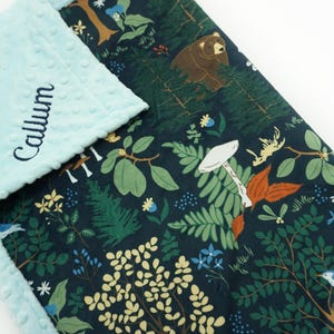 May include: A blue and green baby blanket with a woodland pattern featuring mushrooms, ferns, birds, and a bear. The blanket is personalized with the name "Callum" embroidered on the blue side.