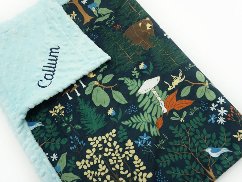 May include: A blue and green baby blanket with a woodland pattern featuring mushrooms, ferns, birds, and a bear. The blanket is personalized with the name "Callum" embroidered on the blue side.