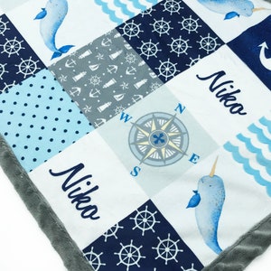 May include: A blue and white baby blanket with a nautical theme. The blanket features a compass, anchors, narwhals, and the name "Nika" embroidered on it.