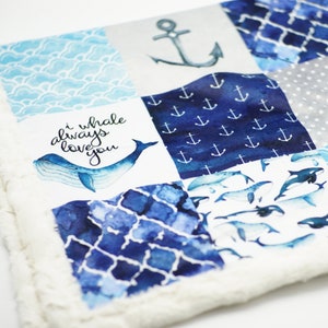 May include: A blue and white baby blanket with a patchwork design. The blanket features a whale with the text "I whale always love you", anchors, and a blue and white wave pattern.