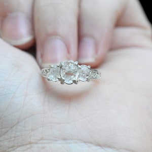 May include: A silver ring with three clear, oval-cut gemstones. The ring is held in a hand against a black background. The gemstones are the focal point, with smaller stones on the sides.