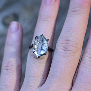 May include: A clear, faceted gemstone ring with a dark metal band. The large, irregular-shaped stone is the focal point, catching the light. The ring is set on a hand, showcasing its size and design. The ring's unique shape and setting give it a modern, artistic look.