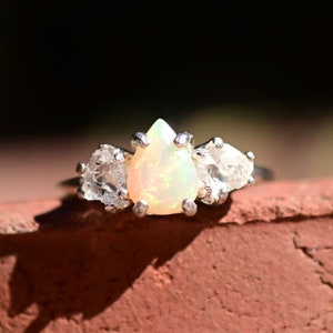 May include: A silver ring featuring a pear-shaped opal gemstone flanked by two smaller, rough-cut gemstones. The opal has a milky, iridescent quality. The ring is set against a blurred, dark background.