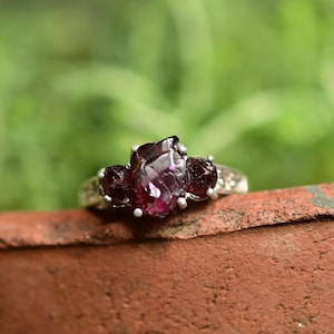 May include: A silver ring featuring three raw, deep red garnet stones set in a prong setting. The central stone is the largest, flanked by two smaller stones on either side, all showcasing natural, irregular facets and a dark, rich crimson hue.