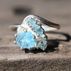 Pode incluir: Um anel de prata com um grupo de pedras preciosas de água-marinha azul claro engastadas em um design delicado em forma de onda.