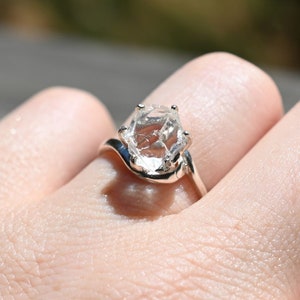 May include: A silver ring with a clear, faceted gemstone set in the center. The ring has a curved band.