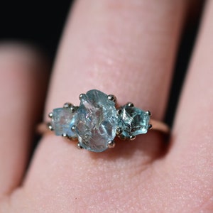 May include: A gold ring with three light blue gemstones set in a cluster. The ring is on a person's finger.