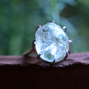 Pode incluir: Um anel de cristal transparente e facetado com uma configuração de prata. O anel está em uma superfície marrom.