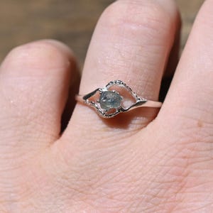 May include: A silver-toned ring featuring a small, round, light blue-grey stone set in a textured, open-work metal frame. The band is thin and polished, with the setting creating a delicate, asymmetrical shape around the central gemstone.