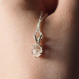 May include: A silver necklace with a small, rough-cut, clear crystal pendant. The pendant is suspended from a delicate chain. The necklace is held in a hand against a soft, neutral background.
