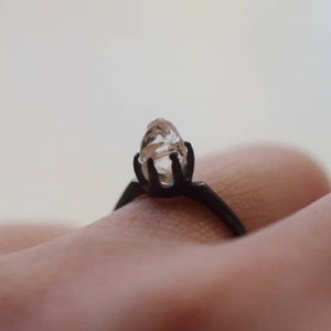 May include: A ring featuring a dark, matte black metal band and a prong setting that holds a single, clear, raw, and irregularly shaped crystal stone. The design showcases a stark contrast between the dark metal and the translucent stone.