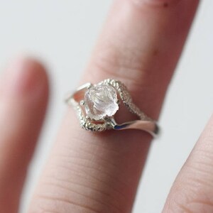 May include: A silver ring with a clear, faceted, raw crystal stone set in the center. The ring has a delicate, curved design.