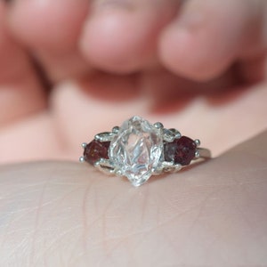 May include: A silver ring featuring a large, clear, marquise-cut central gemstone, flanked by two smaller, round, dark red gemstones. The ring is displayed on an open palm against a dark backdrop. A piece of jewellery.