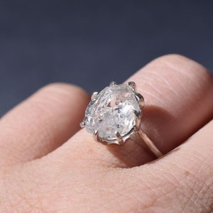 Pode incluir: Um anel de prata com uma grande pedra preciosa clara em forma oval. A pedra preciosa tem uma aparência texturizada e cristalina. O anel é montado em uma simples banda de prata. O anel é mostrado em um dedo.