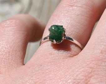 Puede incluir: Un anillo de plata con una gema verde esmeralda en bruto. El anillo tiene una fina banda de plata y la piedra está engastada en un sencillo engaste de cuatro garras. La gema tiene un aspecto natural y sin pulir.