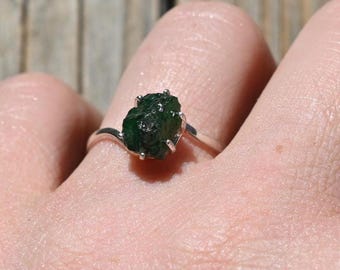 Puede incluir: Un anillo de plata con una gema de esmeralda verde oscuro, en bruto y sin cortar. La esmeralda es el punto focal, engastada en una delicada banda de plata. El anillo se muestra en una mano, mostrando la textura y el color natural de la gema.