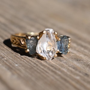 May include: A gold ring with a central pear-shaped clear gemstone flanked by two blue-grey rectangular stones. The ring's band features intricate filigree detailing. The ring is set on a wooden surface.