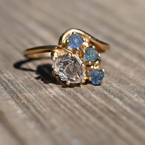 May include: A gold ring with a cluster of gemstones. The ring features a large, clear, faceted stone and several smaller blue stones. The ring is set on a wooden surface, and the sunlight casts a shadow.