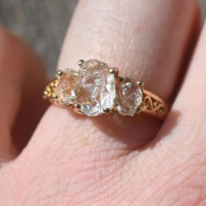 May include: A gold ring with three clear, oval-shaped gemstones. The ring has a detailed filigree design on the band. The gemstones are set close together, creating a cluster effect. The ring is on a person's finger.