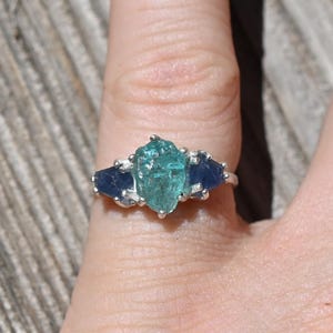 May include: A silver ring with a central light teal gemstone and two dark blue side stones. The ring is displayed on a hand, set against a wooden background. The gemstones' colours provide a striking contrast.