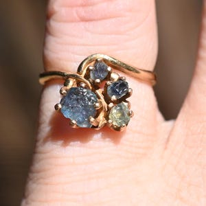 May include: A gold-toned ring featuring a cluster of gemstones in shades of blue and green. The ring's design has a swirling band that leads to the gemstone cluster. The ring is worn on a finger.