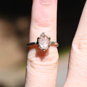 May include: A silver ring with a large, irregularly shaped, clear gemstone. The ring is on a finger, with the hand in the foreground and a blurred outdoor background. The gemstone has a unique, natural appearance.