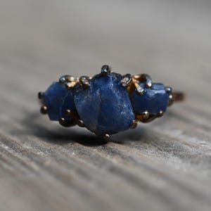 May include: A gold-toned ring featuring three large, irregular-shaped, deep blue gemstones. The ring is set against a blurred, weathered wooden surface, creating a rustic aesthetic. The gemstones are the focal point, catching the light.