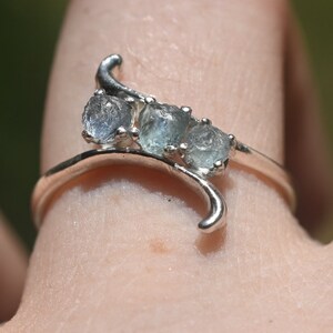 May include: A silver-toned ring featuring a curved, wave-like band design. The ring is set with three small, round, light blue translucent stones arranged in a row along the upper curve of the metal band, which tapers to a point at each end.